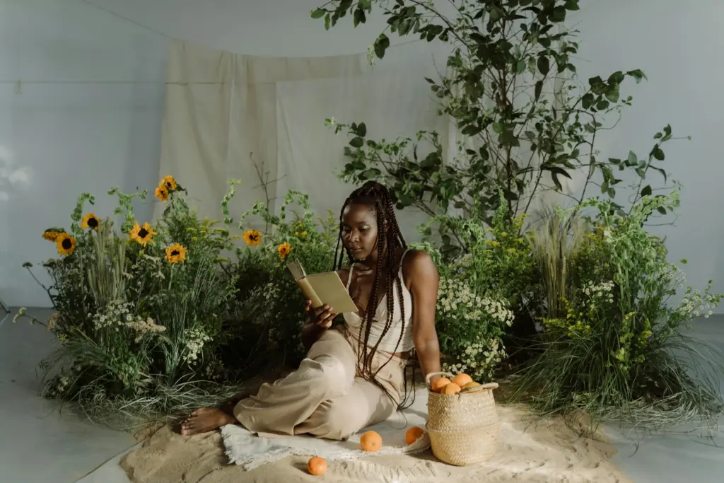 A woman sitting on a blanket surrounded by greenery and sunflowers, reading a book with a basket of oranges beside her, symbolizing relaxation, mindfulness, and natural wellness.